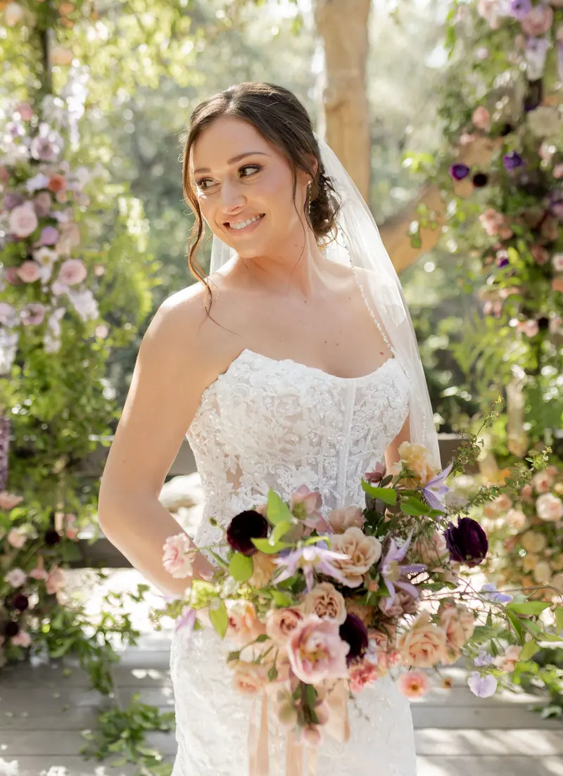 A bride smiling over her shoulder with a pastel bouquet by the flower arch.
