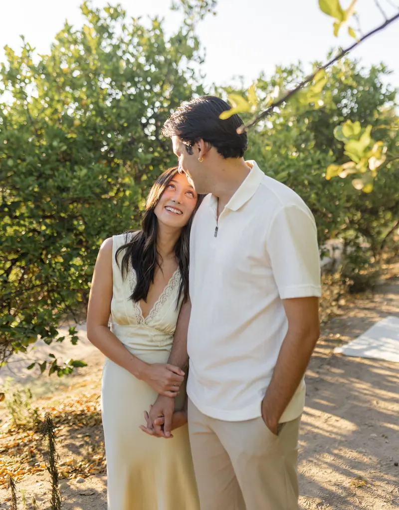 A man kissing his fiancee's forehead in a sunlit grove.