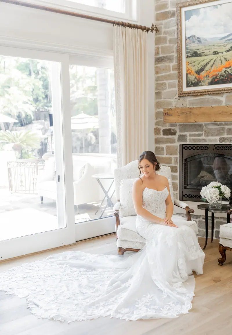A bride seated by a window with her lace train spread across the floor.